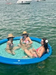 Three women in bikinis and sun hats toast with champagne while lounging in a blue inflatable floating pool on calm green water near anchored boats on a sunny summer boating day.