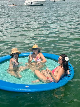 Three women in bikinis and sun hats toast with champagne while lounging in a blue inflatable floating pool on calm green water near anchored boats on a sunny summer boating day.