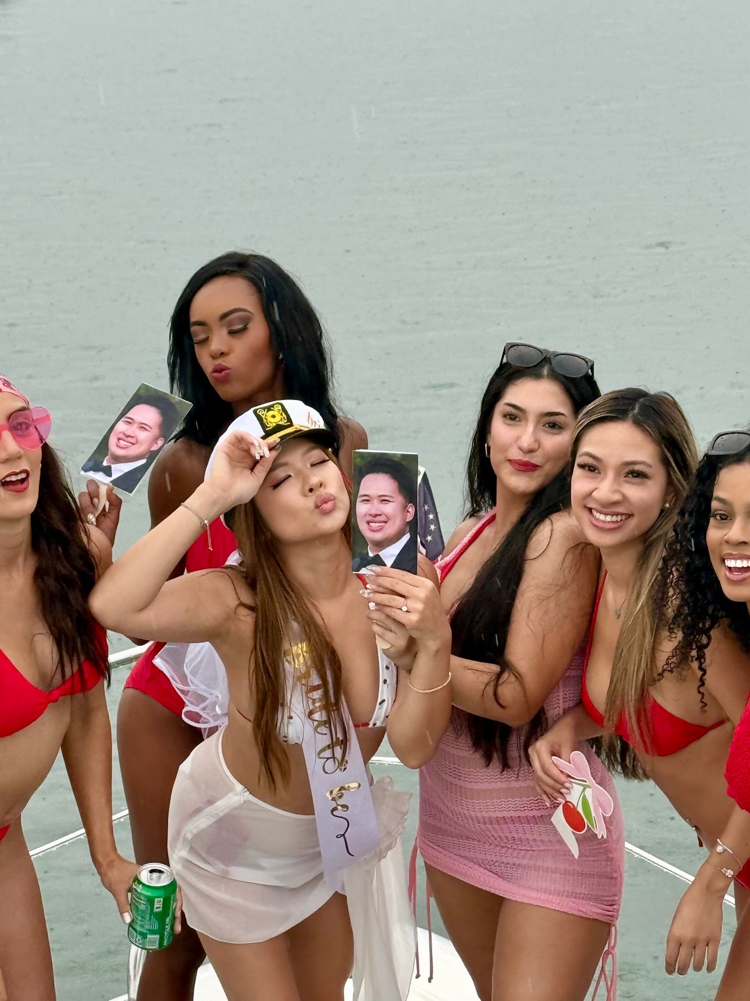 Bachelorette boat party: group of women in swimsuits on a boat deck posing with printed face cutouts; bride-to-be wearing a sash and captain hat, water in the background.