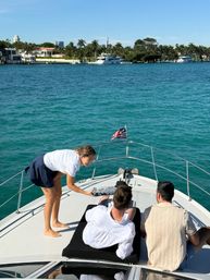 Server handing a tray of drinks to two people lounging on the bow of a yacht in turquoise water near a palm-lined Miami waterfront with luxury boats.