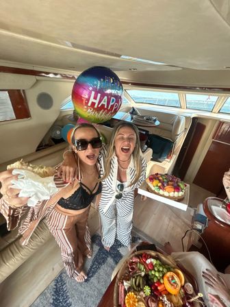 Two women laughing inside a yacht celebrating a birthday with a colorful "Happy Birthday" balloon, fruit‑topped cake, charcuterie board and ocean view through windows.