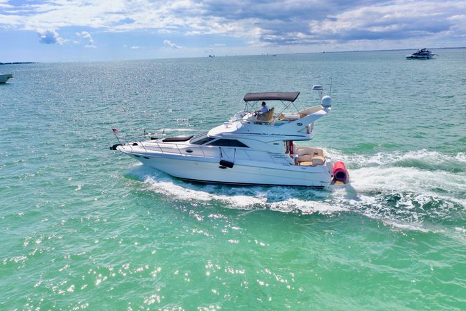 Sunlit white motor yacht cruising through turquoise coastal waters under a partly cloudy blue sky, leaving a foamy wake — vibrant boating seascape.
