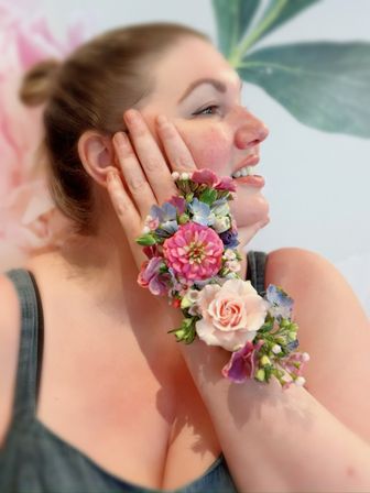 Smiling person with hand on cheek wearing a vibrant wrist corsage of pink, cream, and blue flowers against a soft floral backdrop