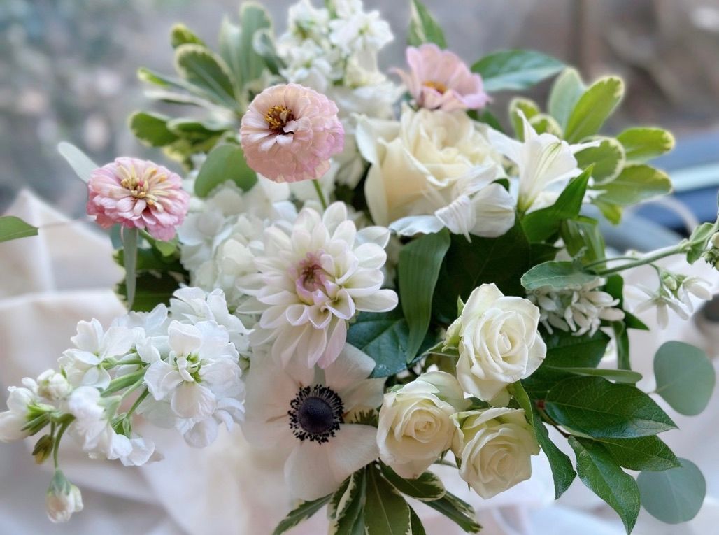 Dreamy close-up of a soft blush-and-white bouquet with roses, dahlia, anemone and mixed greenery resting on draped light fabric