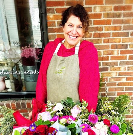 Beaming florist in an apron outside a brick storefront with vibrant mixed bouquets of pink, red, and white blooms
