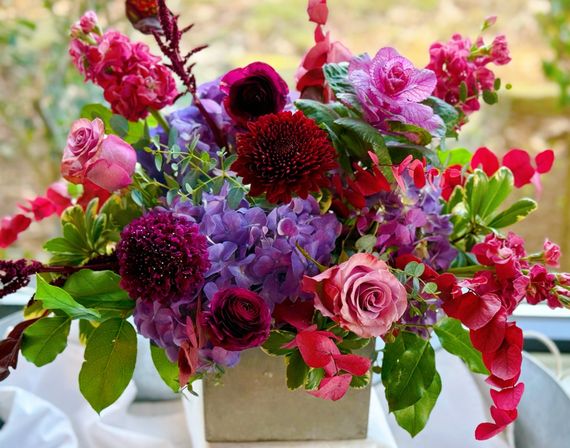 Vibrant pink and purple floral centerpiece with roses, hydrangeas, ranunculus and deep red mums mixed with eucalyptus and green foliage in a square vase on a sunny windowsill