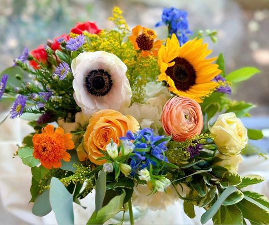 Vibrant garden-style bouquet with sunflower, white anemone, orange ranunculus, yellow rose, blue blooms and lush greenery — bright spring floral centerpiece