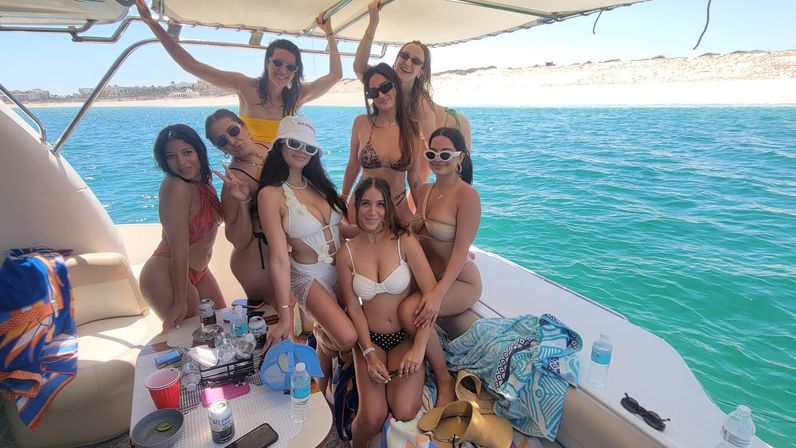 Eight women in bikinis posing on a sun‑shaded boat amid turquoise coastal waters and a sandy beach, with towels and drinks nearby — sunny boat party vibe.
