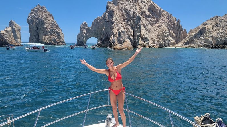 Cheerful woman in a red bikini standing on a boat bow with arms outstretched, turquoise sea and the rocky natural arch of Cabo San Lucas in the background under a clear blue sky.