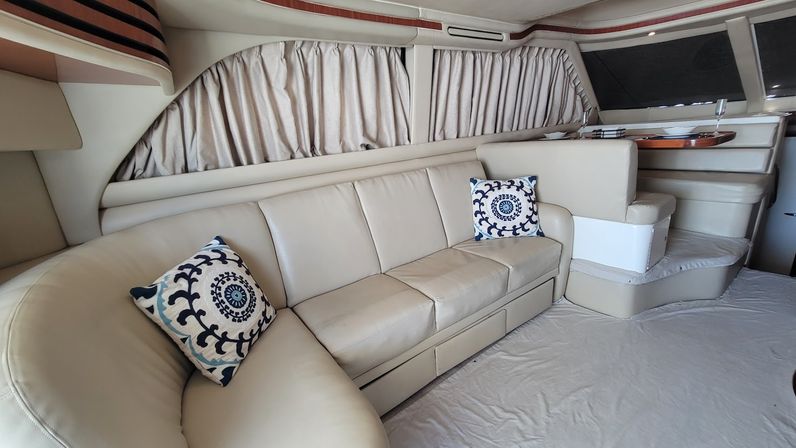 Cozy beige leather curved sofa with two blue patterned throw pillows inside a bright yacht cabin with curtained windows and a small dinette area.