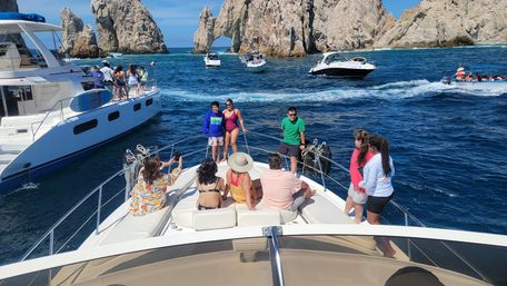 Group of people enjoying the bow of a yacht amid multiple tour boats on deep blue ocean water, with a dramatic rocky sea arch and sunlit cliffs in the background.