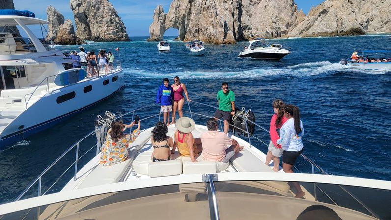 Group of people enjoying the bow of a yacht amid multiple tour boats on deep blue ocean water, with a dramatic rocky sea arch and sunlit cliffs in the background.