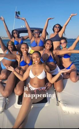Nine women in blue and white bikinis smiling and raising their arms on a sunny boat cruise, ocean and American flag in the background — celebratory boat party vibe.