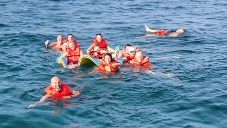 Cheerful group of people in bright orange life jackets floating and smiling around a bodyboard in sunlit blue ocean water.