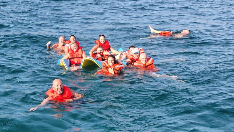 Cheerful group of people in bright orange life jackets floating and smiling around a bodyboard in sunlit blue ocean water.