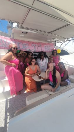 Women in pink outfits posing on a yacht under a "BRIDE TO BE" banner for a sunny bachelorette boat party.