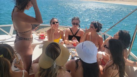 Group of friends in swimsuits gathered around a table on a sunlit yacht near a sandy beach, chatting and sipping drinks over blue coastal waters.