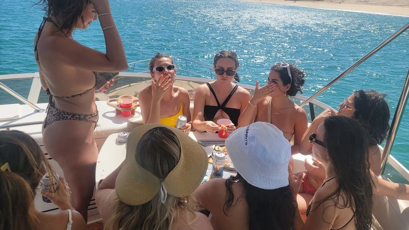 Group of friends in swimsuits gathered around a table on a sunlit yacht near a sandy beach, chatting and sipping drinks over blue coastal waters.