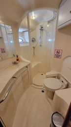 Snug beige yacht bathroom (marine head) with curved vanity and sink, wall-mounted handheld shower in a compact stall, toilet, porthole window, and small trash bin.