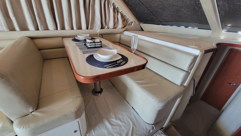 Luxury yacht dining nook with cream leather L-shaped banquette, wood-trim table set for two with white dishes and champagne flutes, curtained windows and built-in storage