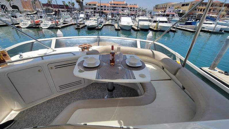 Sunlit yacht dining nook with a small table set for two—wine bottle, glasses and place settings—curved cream leather seating overlooking a busy marina of moored boats and waterfront buildings.