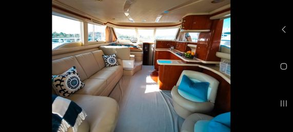 Sunlit luxury yacht salon with cream leather L-shaped sofa, round cream chairs with blue accent pillows, polished wood galley and wraparound windows showing marina views.