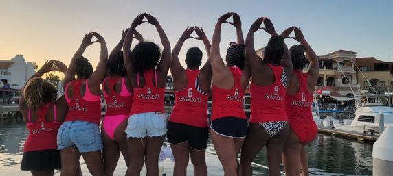 Eight women in matching red tank tops and swimsuits stand with backs to camera at a marina at sunset, arms raised forming heart shapes, docks and boats in the background.