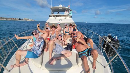 Group of adults lounging on the bow of a motor yacht for a sunny ocean yacht party near a rocky coastline, blue sky and a Mexican flag visible.