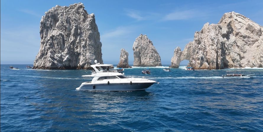Sunlit white motor yacht cruising in deep blue Sea of Cortez waters near the iconic El Arco rock formations at Cabo San Lucas, with smaller tour boats and a clear blue sky.