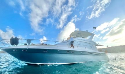 White luxury motor yacht floating on clear turquoise coastal waters under a bright blue sky with fluffy clouds, a person walking along the sunlit deck