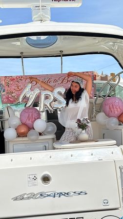Bride-to-be posing on a white yacht at a marina with silver "MRS" balloon, pink "Future Mrs" balloons, floral "Bride to Be" banner and bouquet for a fun bridal shower celebration.