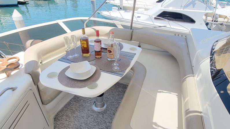 Sunlit yacht dining nook at a marina with curved beige seating and a small white table set with bottles, stemmed glasses and place settings on the boat deck.