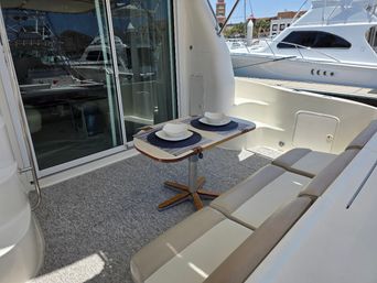 Cozy alfresco dining spot on a white yacht at a sunny marina, small teak table set for two with blue placemats and white bowls, cushioned bench seating and sliding glass door with other boats docked nearby.
