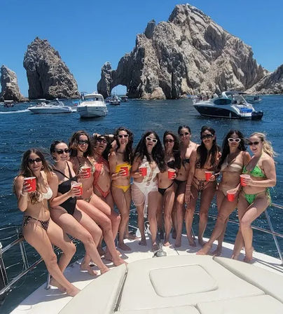 Group of women in bikinis holding red cups on the bow of a yacht, smiling under a bright blue sky with boats and the iconic El Arco rock formations at Cabo San Lucas, Mexico.