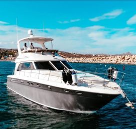 Luxury white motor yacht cruising near a rocky breakwater in a sunny coastal marina, turquoise water and clear blue sky with two people on the flybridge.
