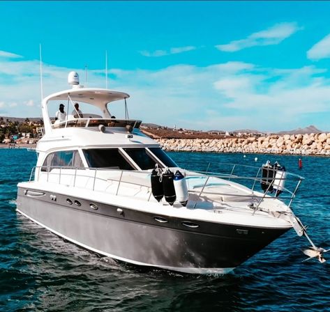 Luxury white motor yacht cruising near a rocky breakwater in a sunny coastal marina, turquoise water and clear blue sky with two people on the flybridge.