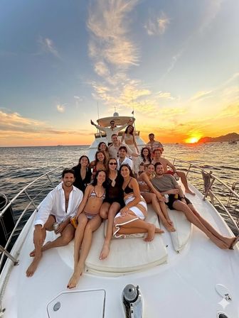 Sunset cruise on a yacht — a smiling group of friends lounging on the bow as the orange sun sets over the ocean and distant coastline, vacation party vibe.