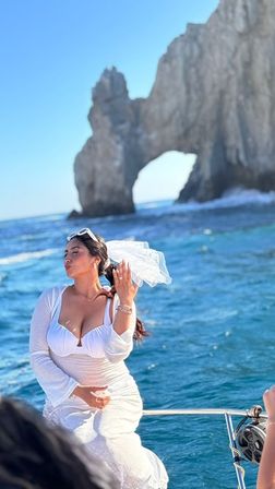 Bride in a white dress and veil playfully posing on a boat near El Arco (Lands End) rock formation in Cabo San Lucas with turquoise sea and clear blue sky