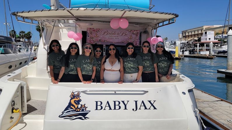 Group of women in matching Cabo shirts and a bride in white posing on a decorated yacht at a sunny marina with pink balloons and a Bride to Be banner, bachelorette vibe.