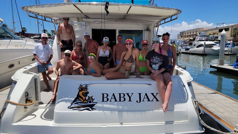 Group of friends in swimsuits posing on the stern of a white motor yacht at a sunny coastal marina, docked beside other yachts with waterfront buildings and a clear blue sky.