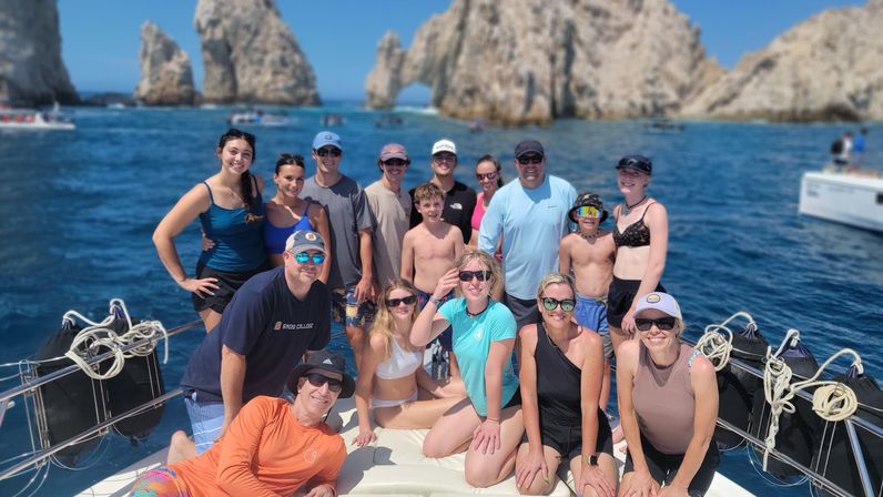 Smiling mixed-age group on a sunny boat tour in turquoise waters near the rocky arches of Cabo San Lucas (El Arco), with other boats nearby.