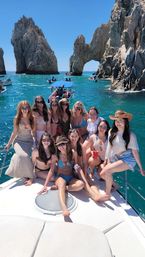 Group of friends on a white boat enjoying a sunny boat party in turquoise waters near a rocky sea arch at Cabo San Lucas, wearing swimsuits, hats, and sunglasses.