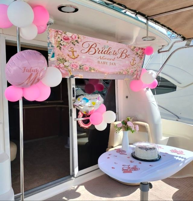 Yacht bridal shower on a boat deck with pink and white balloons, floral “Bride to Be” banner, small table with cake and vase of roses — nautical party decor