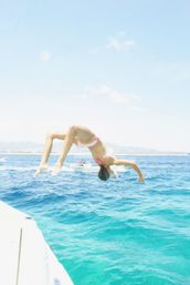 Person mid-backflip off a boat into clear turquoise sea, with distant coastline, other boats and a bright sunny sky