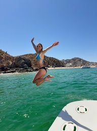 Woman in blue bikini mid-air jumping from a boat into turquoise water near rocky coastline and sandy beach under a clear blue sky