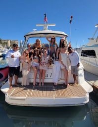 Smiling group of friends and crew posing on the stern of a white yacht at a sunny marina, wearing swimsuits and summer outfits with blue sky, docked boats and harbor buildings in the background — lively yacht party and boat charter scene.