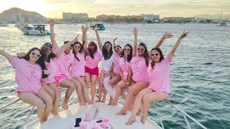 Group of women in matching pink shirts and heart-shaped sunglasses cheering on a yacht at sunset in a busy coastal marina