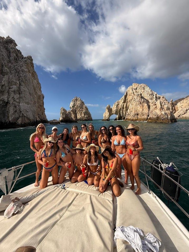 Group of women in colorful bikinis posing and holding red cups on a sailboat near the El Arco rock arch at Cabo San Lucas, with blue ocean and dramatic cliffs beneath a partly cloudy sky.