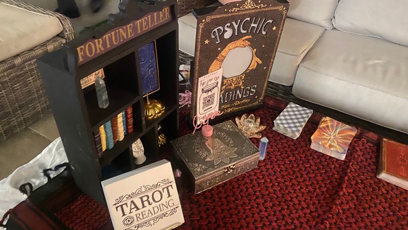 Tarot and psychic reading setup on a red patterned rug in a cozy living room, featuring a fortune teller shelf, psychic readings sign, stacked tarot decks, ornate box and crystal