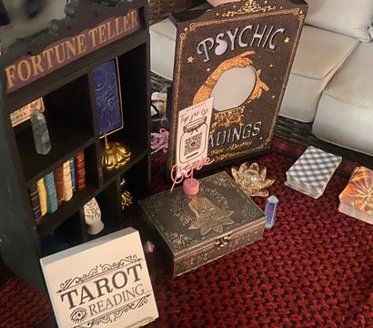 Tarot and psychic reading setup on a red patterned rug in a cozy living room, featuring a fortune teller shelf, psychic readings sign, stacked tarot decks, ornate box and crystal